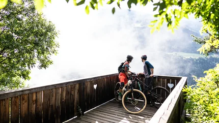 Two mountain bikers on a wooden viewpoint overlooking a foggy valley