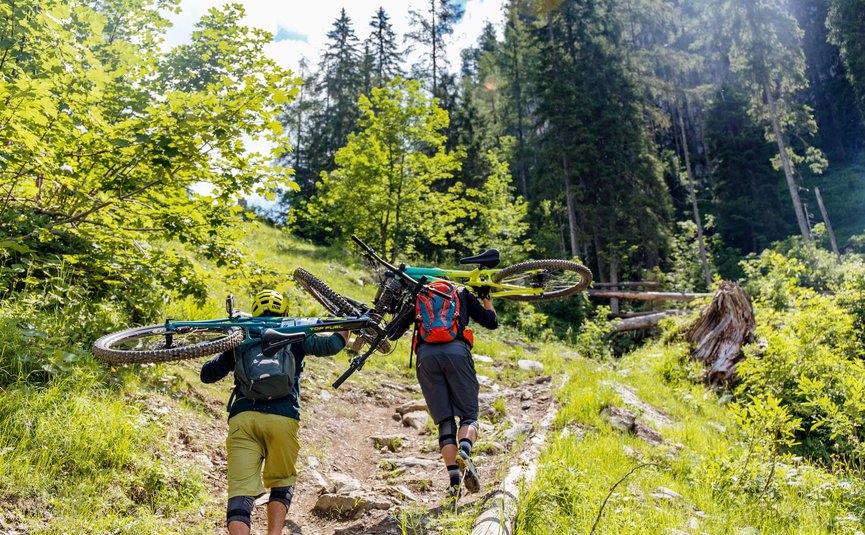 Zwei Mountainbiker tragen ihre Fahrräder auf einem steinigen Waldweg bergauf