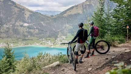 Zwei Mountainbiker blicken auf einen See und Berge von einem Waldpfad aus
