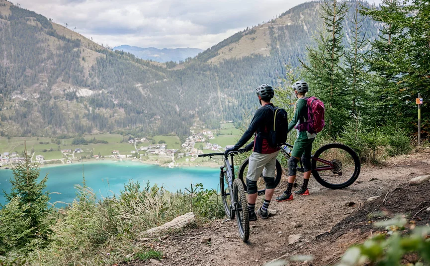 Zwei Mountainbiker blicken auf einen See und Berge von einem Waldpfad aus