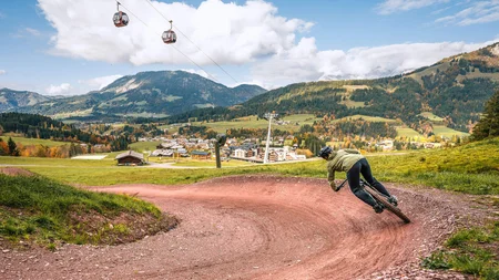 Happy Birthday EPIC Bike Park! © Sebastian Astl Mountainbiker auf kurviger Strecke vor Alpenlandschaft mit Seilbahnen