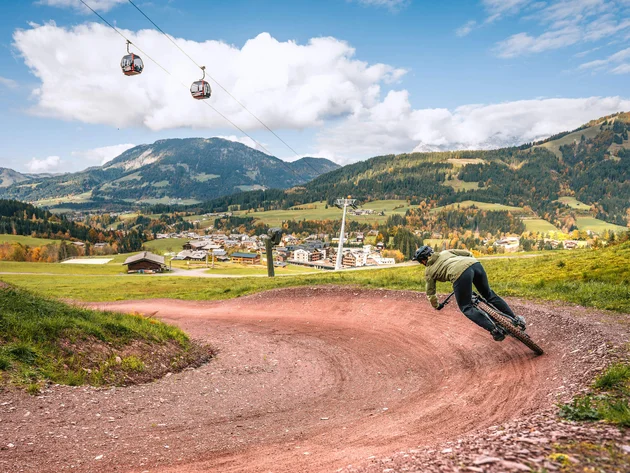 Happy Birthday EPIC Bike Park! © Sebastian Astl Mountainbiker auf kurviger Strecke vor Alpenlandschaft mit Seilbahnen