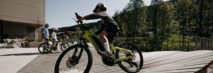 Kind rijdt fiets op houten helling met volwassenen en bomen op achtergrond