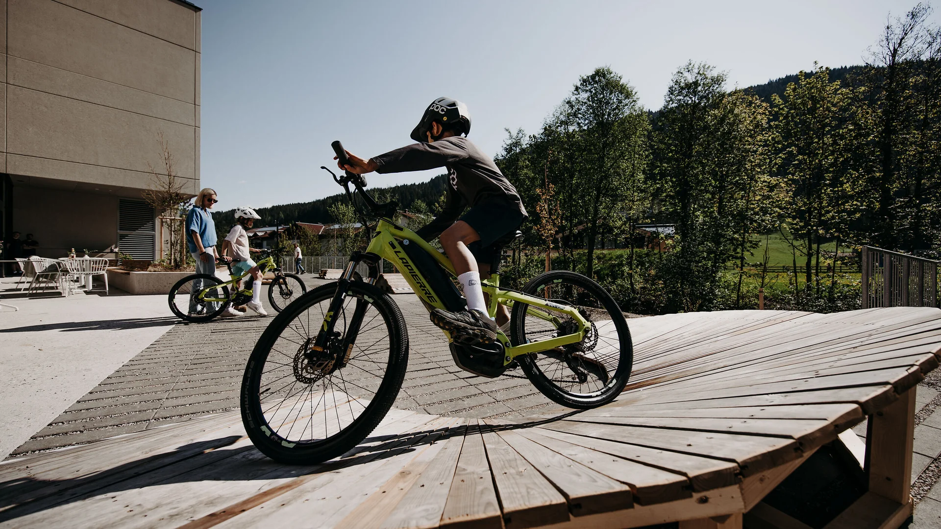 Kind fährt Fahrrad auf Holzrampe mit Begleitern und Bäumen im Hintergrund