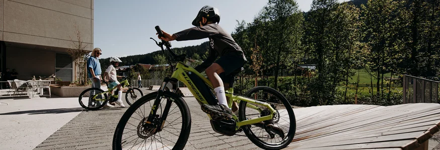 Kind fährt Fahrrad auf Holzrampe mit Begleitern und Bäumen im Hintergrund