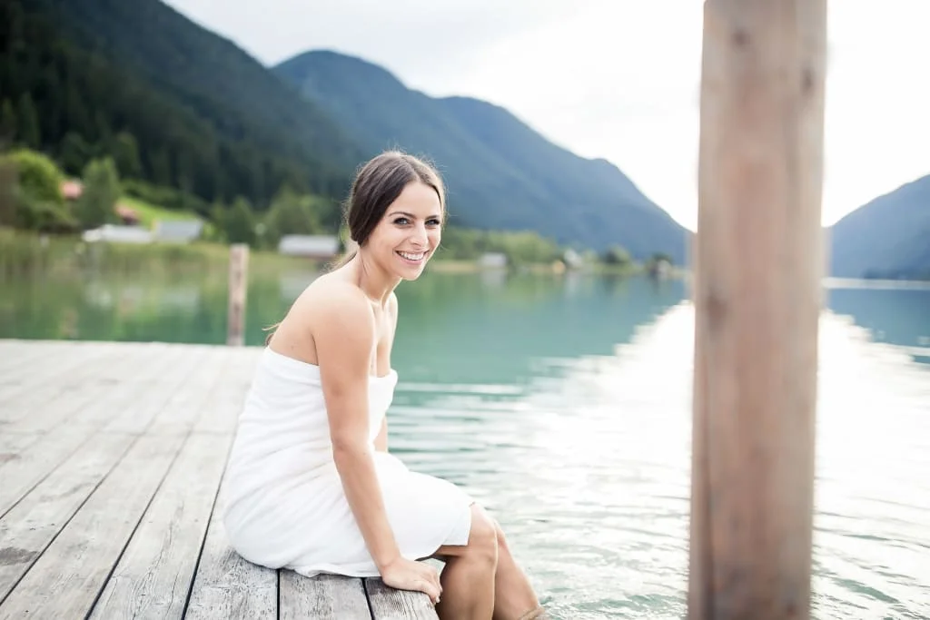 Bewusst Frau Sein Lächelnde Frau im weißen Kleid auf einem Steg an einem Bergsee