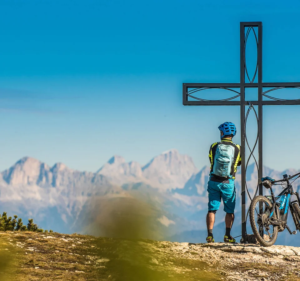 Radfahrer steht neben einem Gipfelkreuz mit Bergpanorama im Hintergrund
