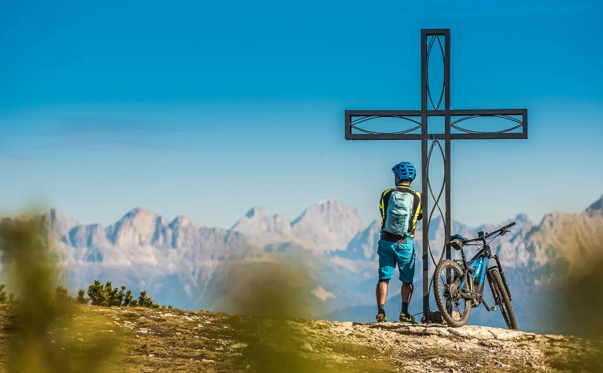 Cyclist standing by summit cross with mountain range in the background