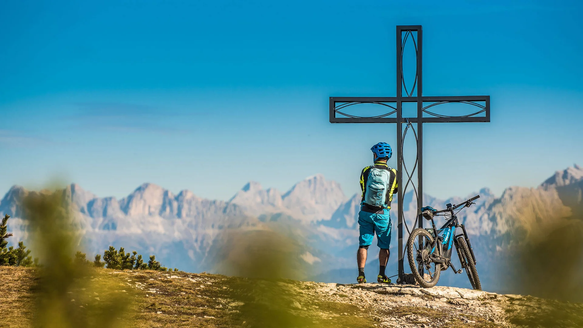 Cyclist standing by summit cross with mountain range in the background