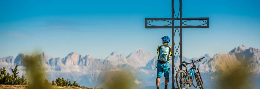 Radfahrer steht neben einem Gipfelkreuz mit Bergpanorama im Hintergrund
