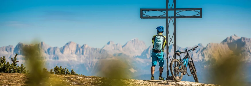 Radfahrer steht neben einem Gipfelkreuz mit Bergpanorama im Hintergrund