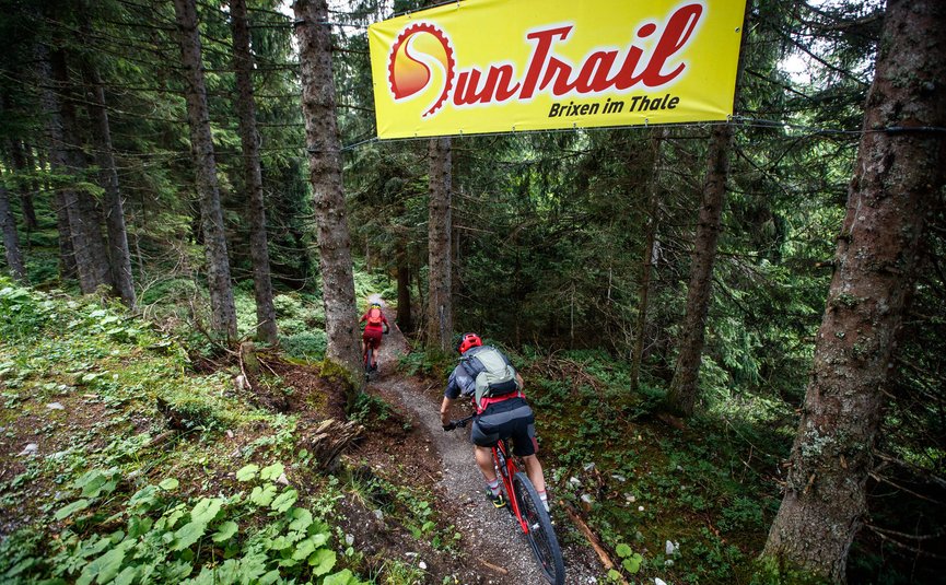 Zwei Mountainbiker fahren auf einem Waldtrail unter einem SunTrail-Banner