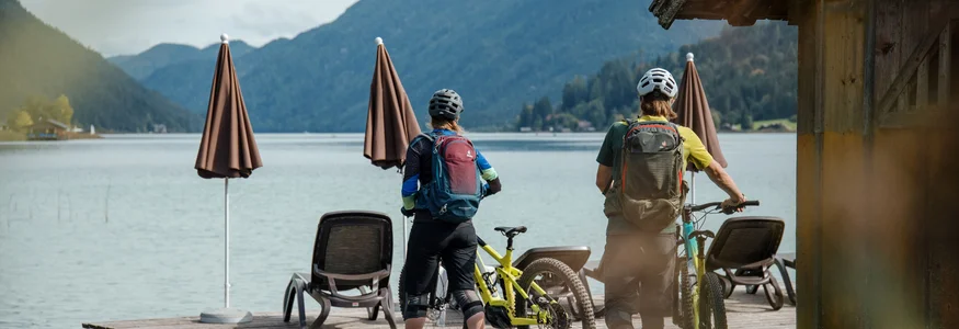 Zwei Radfahrer mit Helmen stehen am See auf einem Steg mit Bergblick
