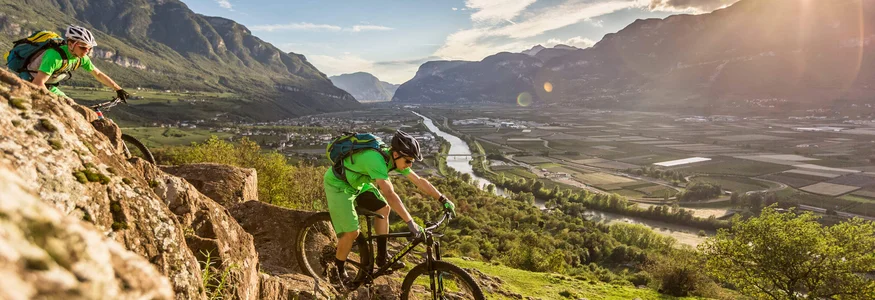 Zwei Mountainbiker fahren auf felsigem Bergpfad mit Tal und Fluss im Hintergrund