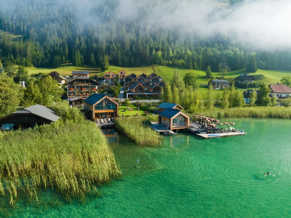 Dorf mit Häusern am klaren See und grünen Hügeln im Nebel