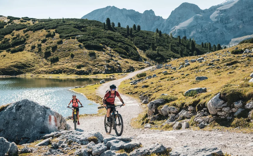 Two mountain bikers riding on a rocky trail beside a lake in the mountains