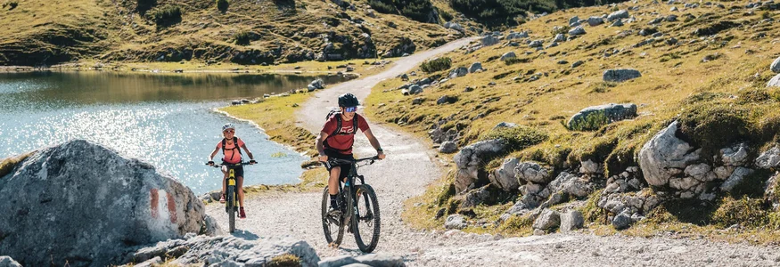 San Vigilio Dolomites - Kronplatz © Alex Moling Zwei Mountainbiker fahren auf einem bergigen Wanderweg neben einem See