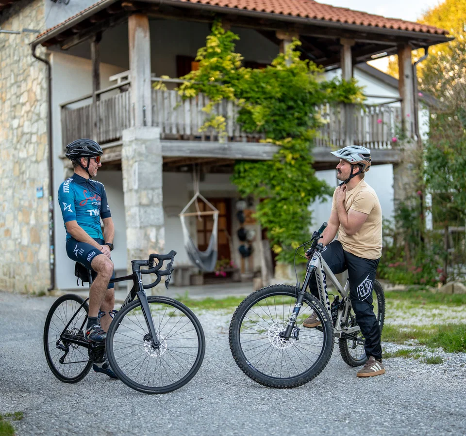 Zwei Männer mit Fahrrädern stehen auf einem Weg vor einem Haus und unterhalten sich