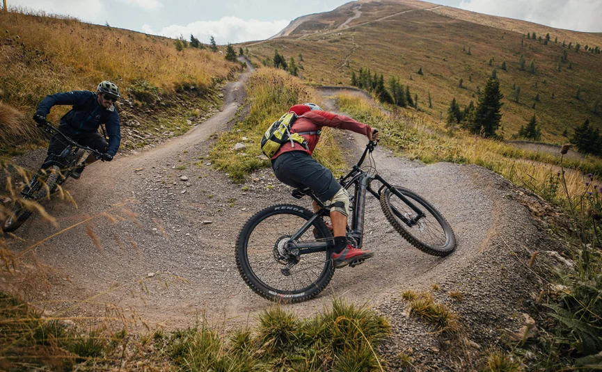 Two mountain bikers riding on a winding mountain trail with grass and trees
