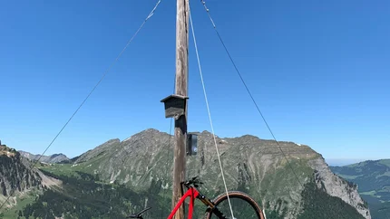 Rotes Mountainbike lehnt an Gipfelkreuz mit Bergblick bei klarem Himmel