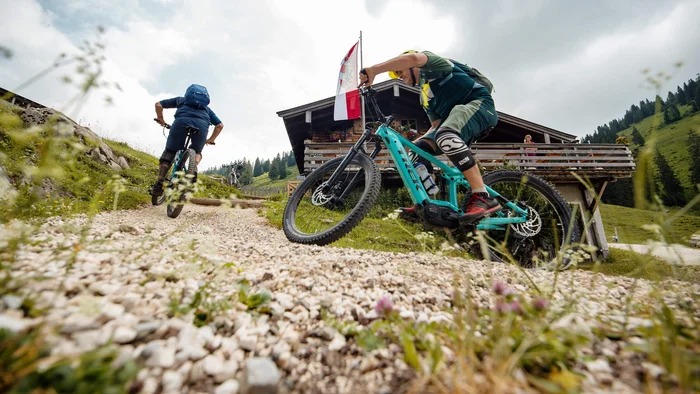 Zwei Mountainbiker fahren auf einem Schotterweg an einer Hütte in den Bergen vorbei