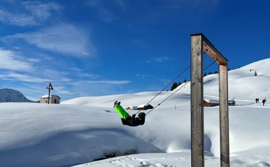 Person schaukelt auf Holzschaukel im verschneiten Bergland unter blauem Himmel