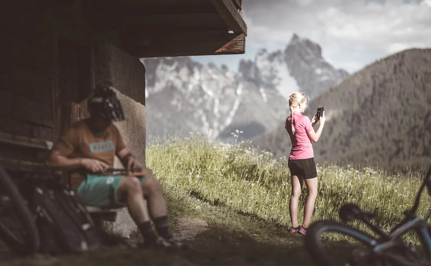 HIRBEN Naturlaub **** © Manuel Kottersteger Woman taking photo of mountain, man with helmet sitting on bench by bike