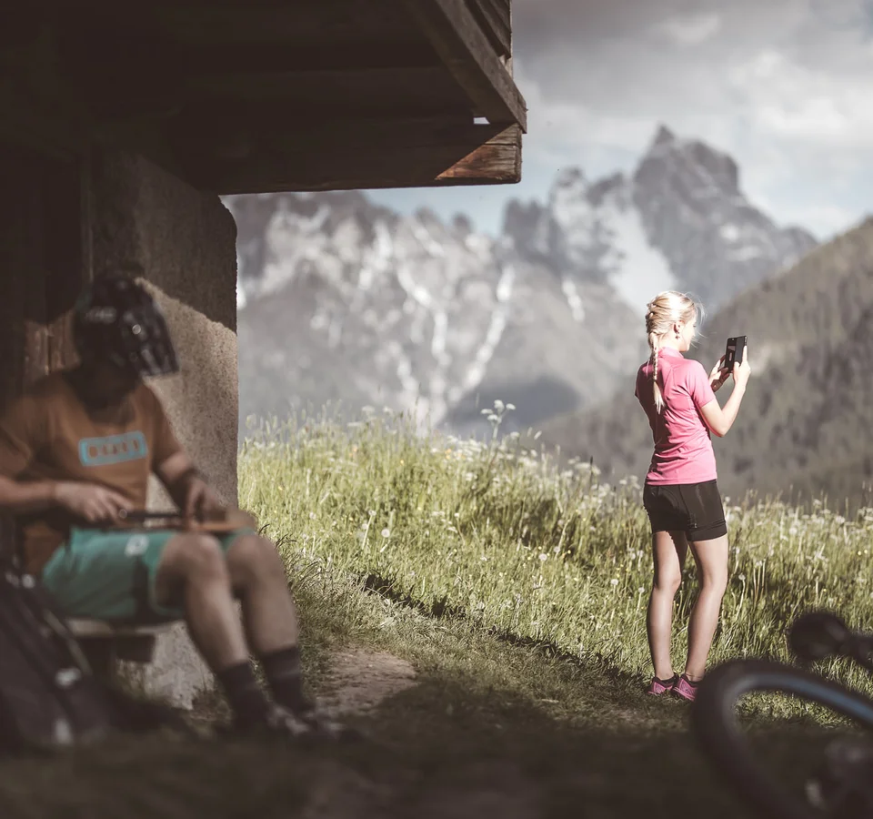 Frau fotografiert Berglandschaft, Mann mit Helm sitzt auf Bank neben Fahrrad