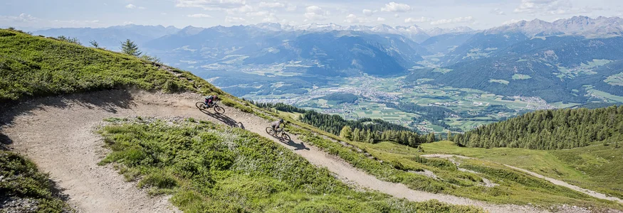 Zwei Mountainbiker fahren auf einem Bergpfad mit Alpenpanorama