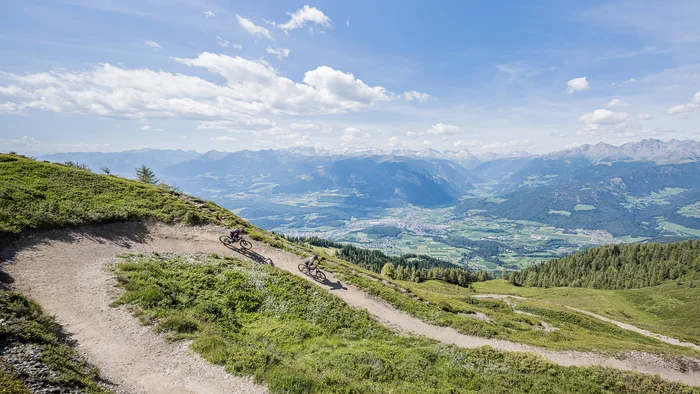 Zwei Mountainbiker fahren auf einem Bergpfad mit Alpenpanorama