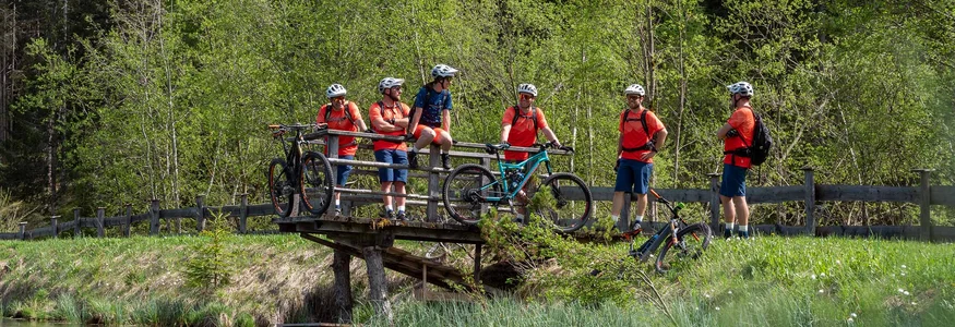 Gruppe von Radfahrern in orangefarbenen Trikots auf einer Holzbrücke neben einem Teich