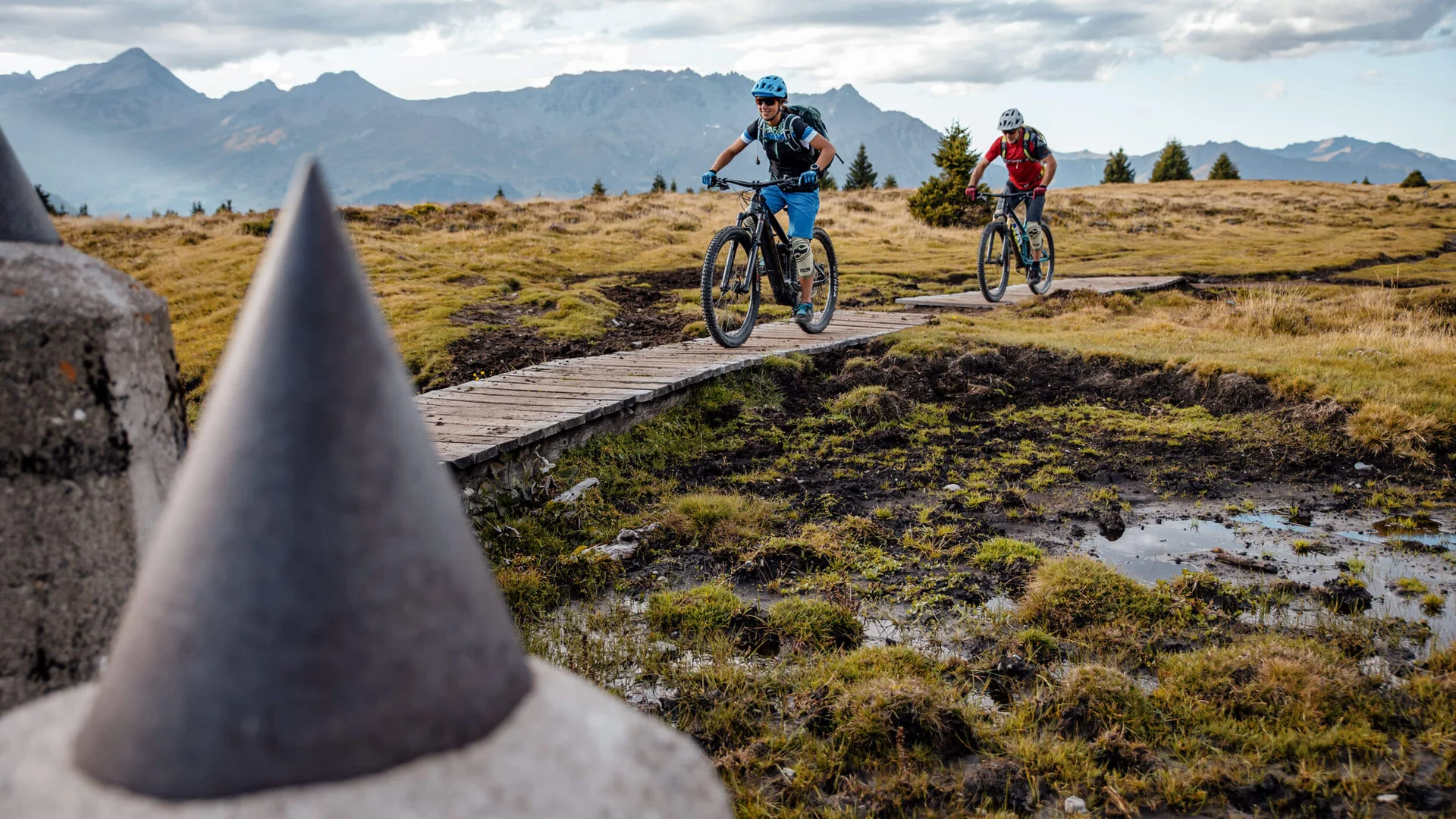Zwei Mountainbiker fahren auf einem Holzsteg durch Gebirgslandschaft