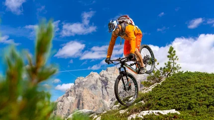 Mountain biker in orange gear jumps on rocky alpine trail under sunny sky