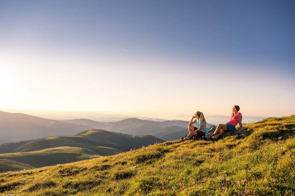 Two women sitting on a grassy hill overlooking mountains at sunset
