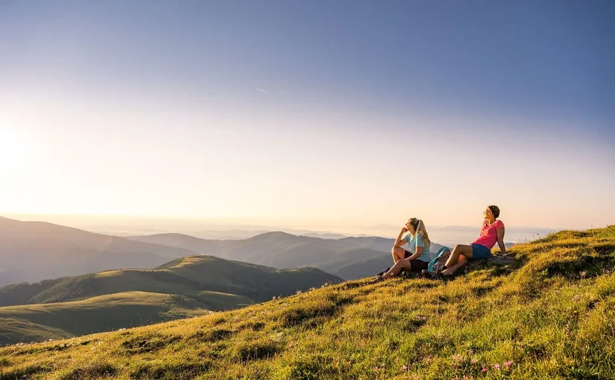 Two women sitting on a grassy hill overlooking mountains at sunset