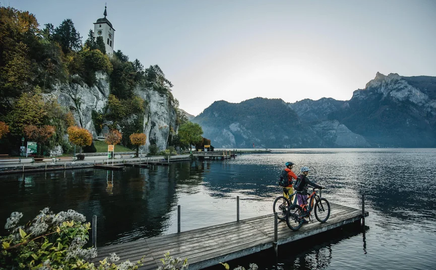 Twee fietsers op een steiger bij een meer met bergen en kerk op de achtergrond