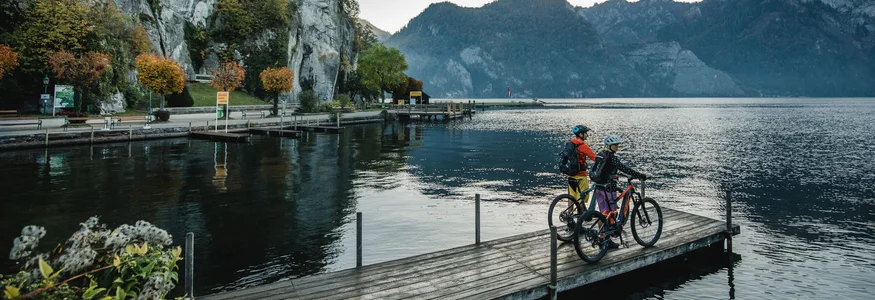 Mountainbikevakantie in Opper-Oostenrijk © Salzkammergut Tourismus - Katrin Kerschbaumer Twee fietsers op een steiger bij een meer met bergen en kerk op de achtergrond