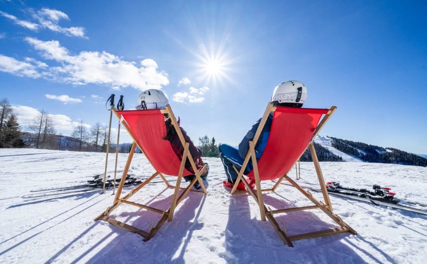 Short getaway with ski or therme Two skiers sitting on red deck chairs in snowy landscape under bright sun