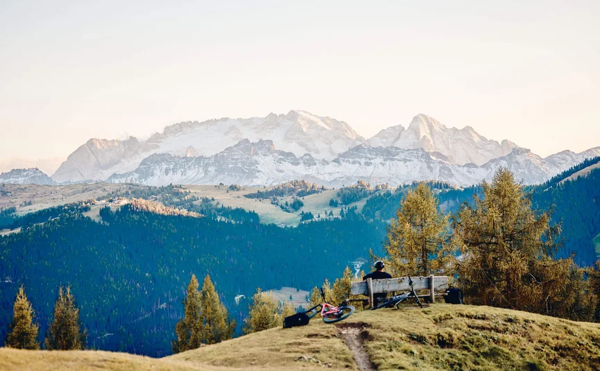 Person sitzt auf Bank, Blick auf bewaldete Hügel und schneebedeckte Berge