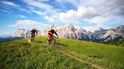 Zwei Mountainbiker fahren auf einem Pfad durch eine grüne Bergwiese vor Felsbergen