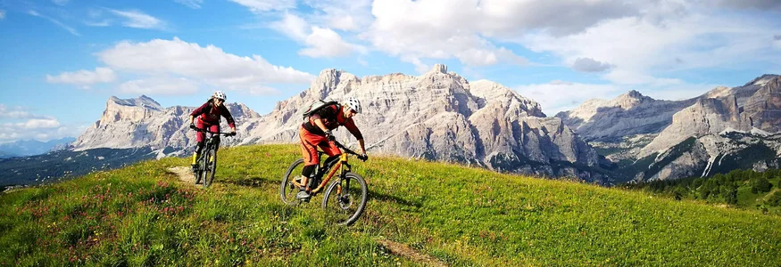Zwei Mountainbiker fahren auf einem Pfad durch eine grüne Bergwiese vor Felsbergen