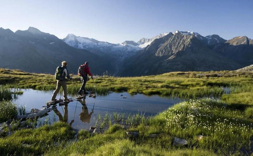 Zwei Wanderer überqueren einen kleinen Bach in einer bergigen Landschaft