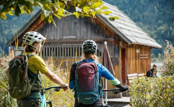 Gradys Gravel Queens Event 2027 Twee fietsers met helmen en rugzakken bij een houten huis in de natuur