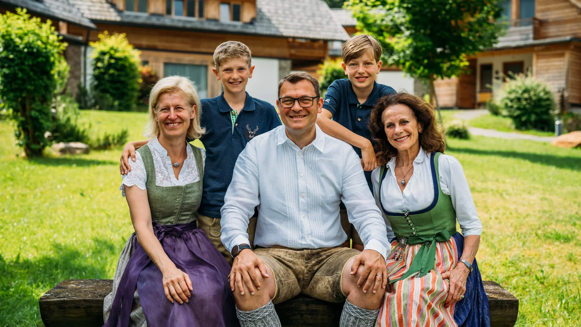 Fünf lächelnde Personen in Trachten vor Holzhäusern im Grünen