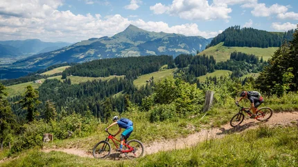 Zwei Mountainbiker fahren auf einem Bergweg mit grüner Landschaft und Bergpanorama