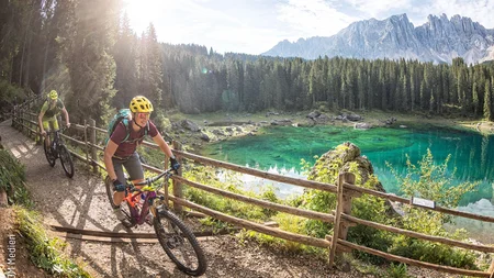 Two cyclists with helmets riding on a forest path by a clear mountain lake