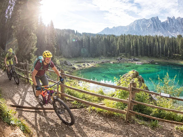 Two cyclists with helmets riding on a forest path by a clear mountain lake