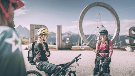 Zwei Mountainbikerinnen mit Helm vor großem ROS-Schild in den Bergen