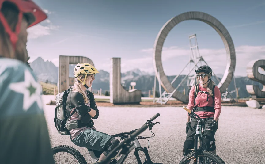 Zwei Mountainbikerinnen mit Helm vor großem ROS-Schild in den Bergen