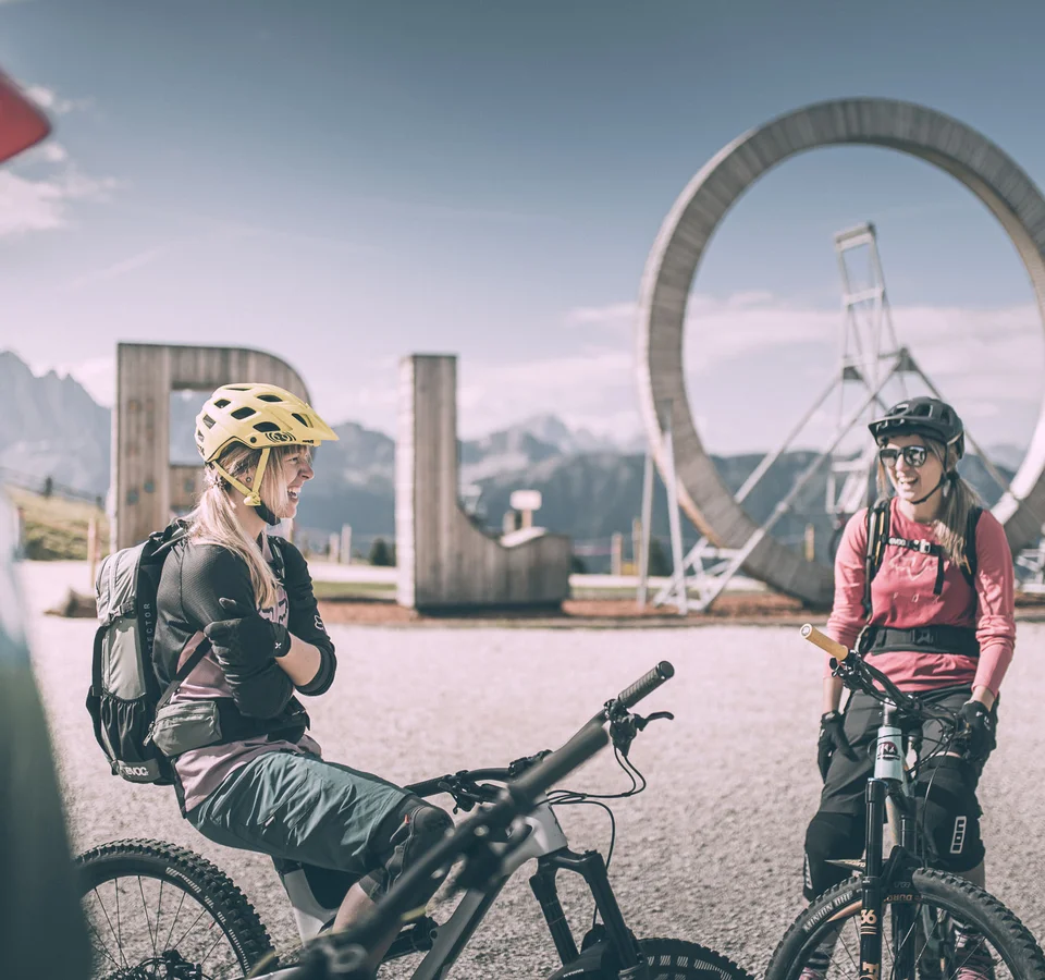 Biken á la Brixen © Brixen Tourismus - Manuel Kottersteger Zwei Mountainbikerinnen mit Helm vor großem ROS-Schild in den Bergen